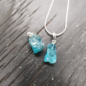 Two Apatite rough pendants - translucent blue/green stone with a rough texture - on a silver chain, on a dark wooden board