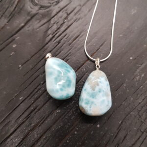 Two Larimar pendants - pale blue with thick white veining - on a silver chain, on a dark wooden board