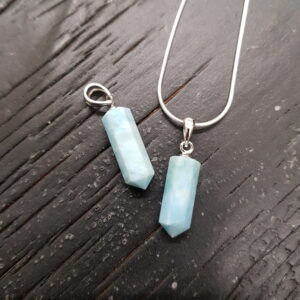 Two Larimar Tritube pendants - three sides tube with rounded edges in a pale blue stone - on a silver chain, on a dark wooden board