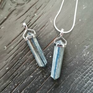 Two Kyanite Double Terminated points (pale blue facets with points at both ends), on a silver chain, on a dark wooden board