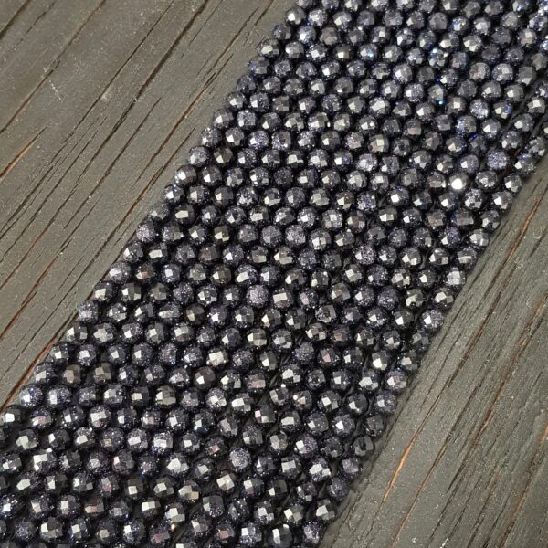 Close up of Blue Goldstone Faceted 4mm beads - rows of small angular dark blue with copper flecked beads on a dark wooden board