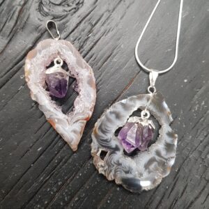 Two Geode Slice with Amethyst Pendants - cream or grey circular agate, with a natural purple amethyst point suspended inside - on a dark wooden board