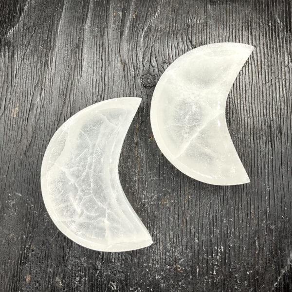 Two Selenite Moon Bowls from above - translucent white stone carved into the shape of a crescent moon - on a black wooden board