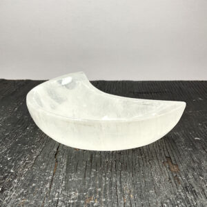 One Selenite Moon Bowl from the side - translucent white stone carved into the shape of a crescent moon - on a black wooden board